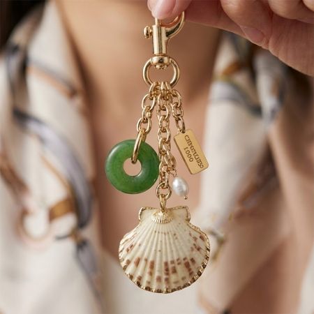 Hand, die einen luxuriösen goldfarbenen Taschenanhänger mit einer Jakobsmuschel, einem grünen Jade-Donut-Anhänger, einer Perle und einem individuellen Metallschild hält.