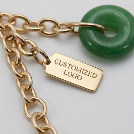 Makro-Detail des gravierten Metallschilds mit individuellem Logo, goldener Kettenglied und poliertem grünen Jade-Anhänger für Modeaccessoires.
