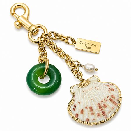 Nahaufnahme des hochwertigen goldfarbenen Taschenanhängers mit individuellem Metallschild, Muschel, Jade-Ring und Süßwasserperle.