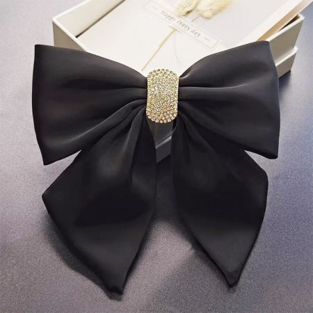 El clip para el cabello con lazo negro con su cierre de piedras de strass doradas, presentado en una elegante caja de regalo blanca.