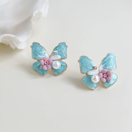 Toma estilizada de los pendientes de mariposa azul junto a flores rosas, destacando los acentos de perlas.