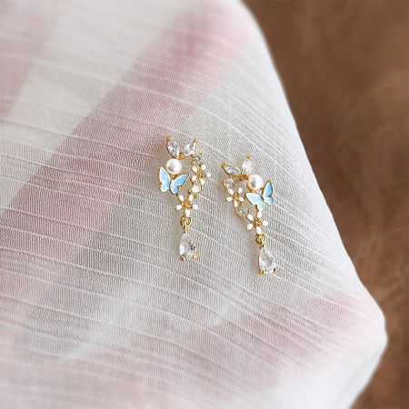 Boucles d'oreilles en émail en forme de papillon et de fleur avec perles zircon, plaquées or 14k en gros. - Une paire de boucles d'oreilles plaquées or 14k avec un papillon en émail bleu détaillé et une fleur blanche, accentuée par une perle pendante.