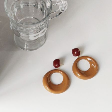 円形のトップとダークレッドのスクエアストーンアクセントを特徴とする、光沢のある茶色のイヤリングの詳細ビュー。