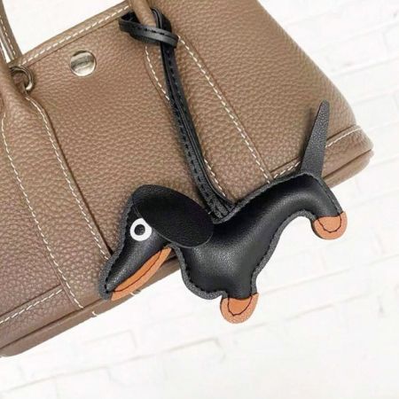Un charm de cuero marrón de dachshund adjunto al mango de un bolso de cuero marrón a juego.