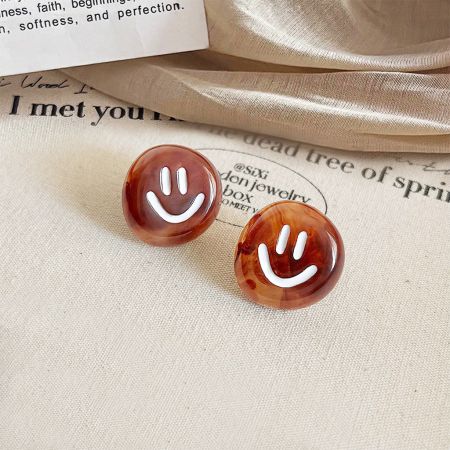 Un paio di orecchini a forma di faccina sorridente marrone esposti su uno sfondo di tessuto.
