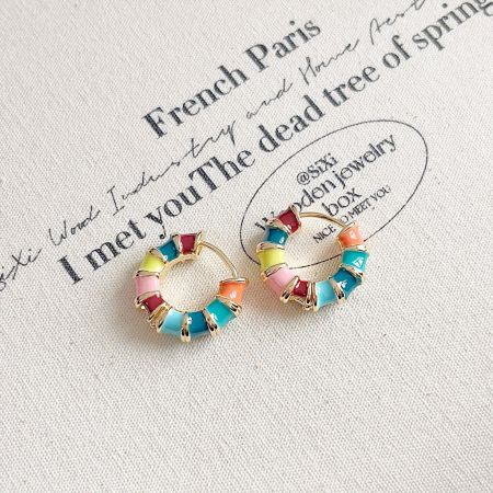 A pair of rainbow enamel bamboo hoop earrings with a gold setting, shown on a branded fabric background.