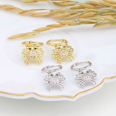 Un paio di orecchini a cerchio huggie con granchio in oro e argento esposti su una superficie decorativa.