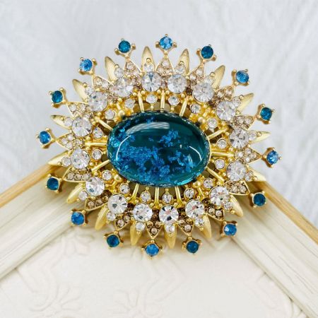 Close-up van de gouden zonnestraal broche met de ingewikkelde details van de centrale blauwe edelsteen en omliggende kristallen.