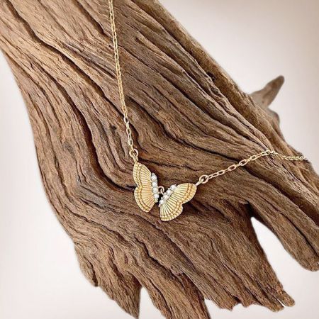 Le collier en or en forme de papillon avec des ailes texturées exposé sur un morceau de bois flotté.