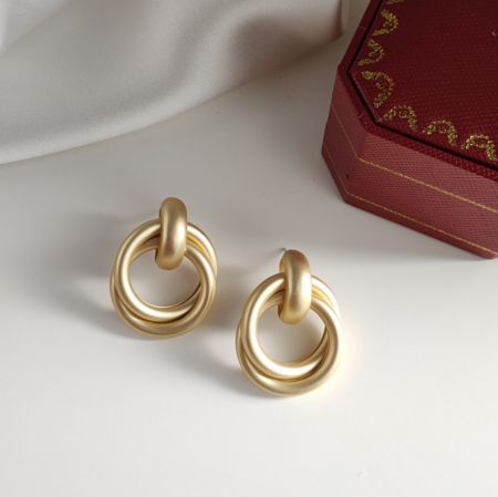 Los pendientes de doble anillo en oro mate exhibidos junto a una vibrante caja de joyería roja.