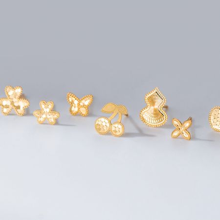Conjunto de pendientes de botón cortados con láser de plata esterlina 925 al por mayor. - Una colección de cinco pares de pendientes de botón cortados con láser de plata esterlina 925 al por mayor en oro, con diseños de flor, mariposa, corazón, cereza y calabaza, dispuestos sobre un fondo blanco.