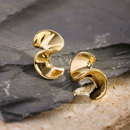 Zijprofiel van de gouden geometrische golf oorbellen tegen een donkere leisteen achtergrond.