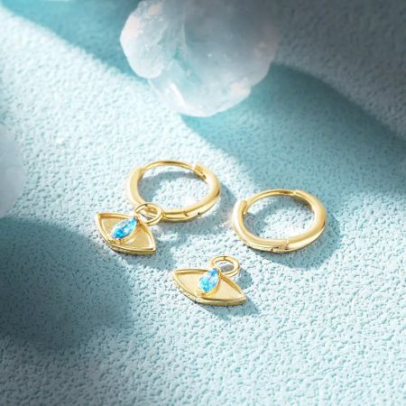 Un paio di orecchini huggie in oro, uno con un ciondolo di occhio maligno pendente.