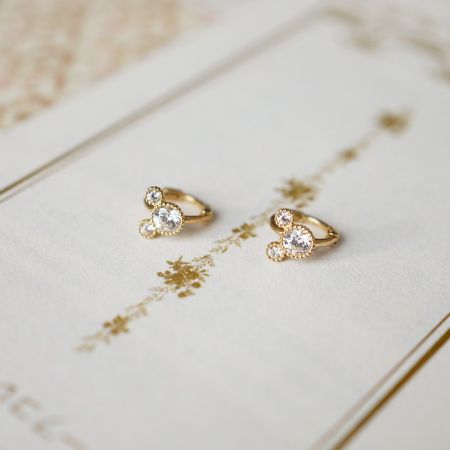 Um par de brincos huggie de prata esterlina com pedras CZ claras em um suporte triangular, exibidos em um fundo branco decorativo.