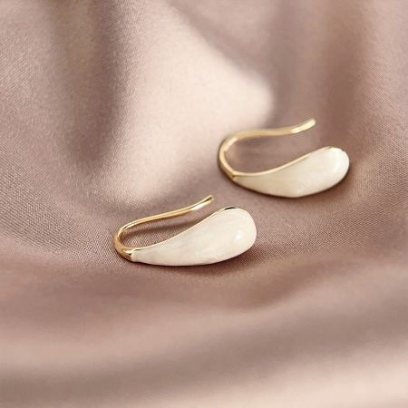 Boucles d'oreilles à crochet français en gros plaquées or avec émail blanc en forme de goutte d'eau - Une paire de boucles d'oreilles à crochet français plaquées or en gros avec des pendentifs en émail blanc en forme de goutte d'eau, présentée sur une surface beige texturée.