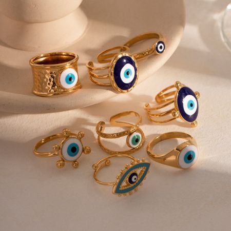 Uma visão de perto de vários anéis de olho grego em aço inoxidável banhados a ouro, exibindo diferentes designs.