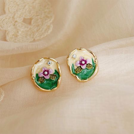 クリーム色の布の背景にピンクの花と緑の葉が描かれた金縁の花のイヤリングの詳細ビュー。