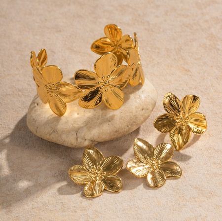 Conjunto de flores de acero inoxidable - Conjunto de joyería de flores de acero inoxidable elegante con chapado en oro, que presenta pendientes y pulsera a juego exhibidos sobre una piedra decorativa.