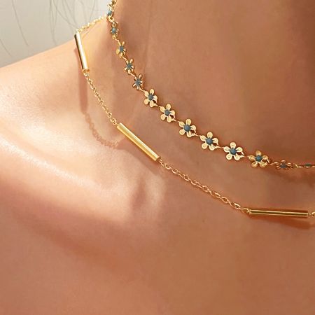 Primo piano della collana dorata con fiori di camomilla in smalto blu esposta su una superficie neutra.
