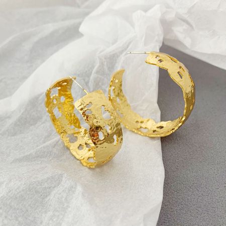 Un paio di orecchini a cerchio perforati in oro con un'estetica unica e fatta a mano su uno sfondo neutro.