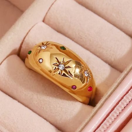 Un anillo de explosión estelar dorado con piedras preciosas coloridas exhibido en una caja de joyería rosa.