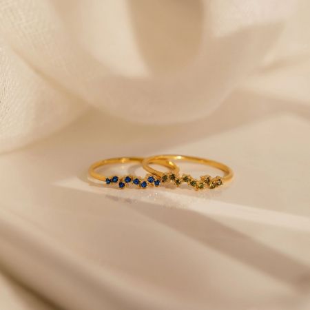 Two stackable birthstone rings, one with blue and one with green gemstones, displayed on a neutral background.
