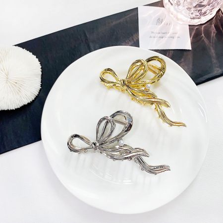 Pinzas para el cabello de cinta grande de metal con acento de perla - Un par de elegantes clips para el cabello en forma de lazo de metal, uno en un acabado dorado pulido y el otro en un acabado plateado elegante, exhibidos sobre una superficie blanca.