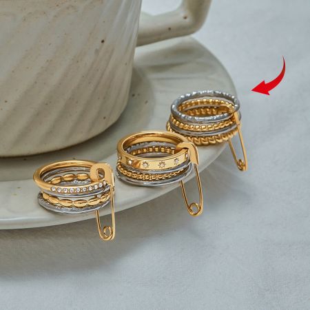 O conjunto completo de cinco anéis empilháveis de ouro e prata, incluindo o anel com detalhe de alfinete de segurança, exibido em um fundo neutro.
