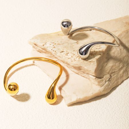 Roestvrijstalen Curve asymmetrische manchetarmbanden met opening, set van 5 - Een set van twee roestvrijstalen cuff bangles, één goud en één zilver, met een modern asymmetrisch druppelontwerp, weergegeven op een gestructureerd stenen oppervlak.