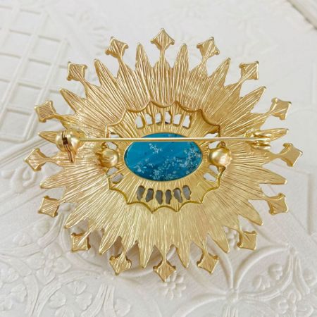 Alternatief ontwerp van de zonnestraal broche met een centrale turquoise steen in een goudkleurig frame.
