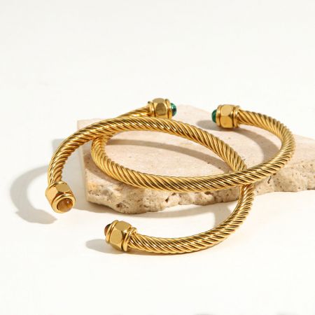 Tre staplade armband i rostfritt stål med vriden design och ädelstensändar, visade på en beige sten.