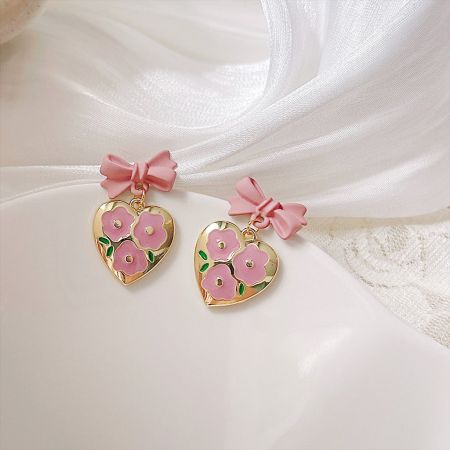 エナメルの花が付いたピンクリボンハートの金色ハートペンダントドロップイヤリング（5ペア） - エナメルの花のディテールが施されたピンクリボンハートドロップイヤリングのペアで、白い背景に展示されています。金色のハートペンダントの上には繊細なピンクのリボンがあります。