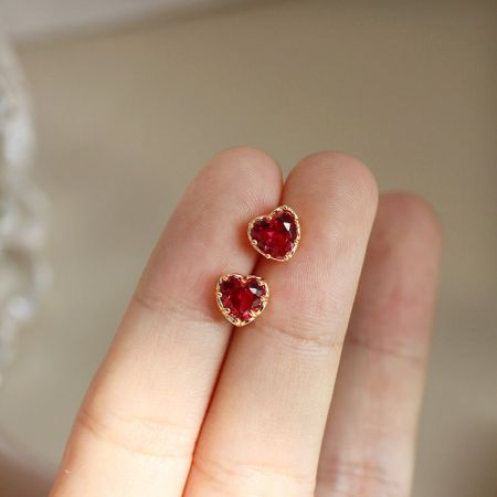 Orecchini a perno a forma di cuore rosso placcati in oro in argento sterling 925 (5 paia) - Un primo piano di delicati orecchini a perno in argento sterling 925, con un design a cuore rosso placcato in oro, tenuti tra le dita per mostrare la scala.