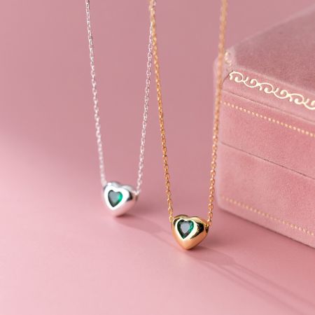 Un par de collares de corazón en acabados de oro y plata, con vibrantes piedras preciosas verdes.