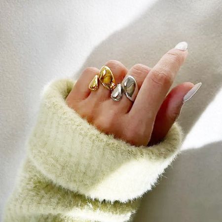 Um close-up de três anéis assimétricos inspirados em folhas em uma mão, disponíveis em acabamentos de ouro e prata.