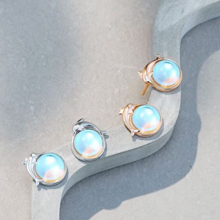 Un assortimento di orecchini a forma di delfino in argento e oro rosa con pietra di luna esposti su una superficie bianca.