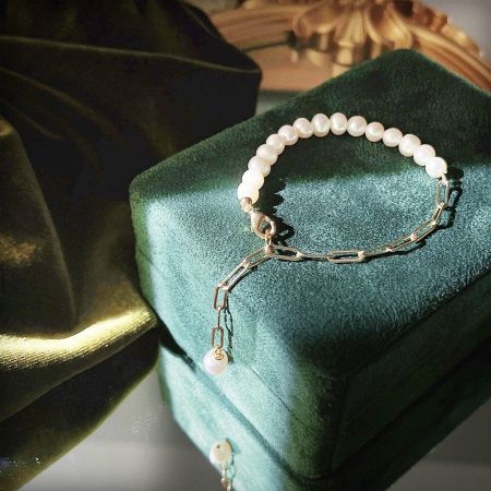A delicate pearl bracelet with a gold paper clip chain and charm, displayed inside a green velvet jewelry box.