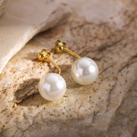 Pendientes de perlas individuales de estilo francés de acero inoxidable - Un par de pendientes de perlas individuales de estilo francés clásico con postes de oro, elegantemente exhibidos sobre un fondo de piedra texturizada.