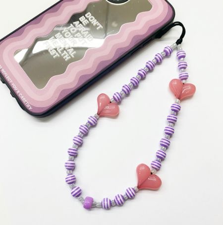 Correa de charm para teléfono con cuentas en forma de corazón. - Correa de charm para teléfono con cuentas en forma de corazón, con cuentas a rayas coloridas y charms en forma de corazón, exhibida junto a una funda de teléfono transparente para mostrar su uso como un accesorio elegante.