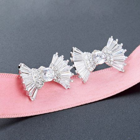 Gli orecchini a forma di fiocco in argento con gemme trasparenti elegantemente presentati su un nastro rosa.
