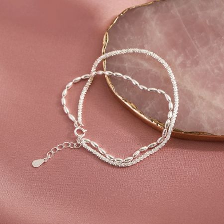 925 Sterling Zilveren Verstelbare Dubbele Laag Kettingarmband - Een close-up van de 925 Sterling Zilveren Verstelbare Dubbele Laag Kettingarmband, die het delicate dubbele kettingontwerp op een zachte roze achtergrond toont.