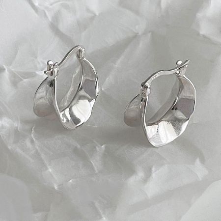 Primo piano degli orecchini a cerchio larghi a onda in argento sterling su una superficie bianca testurizzata.