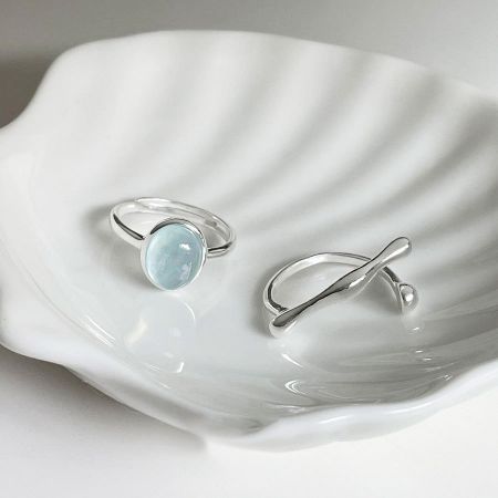 Set di anelli ovali in argento sterling 925 con pietra blu e anelli irregolari regolabili - Un set di anelli in argento sterling 925 regolabili, uno con una pietra azzurra e l'altro con una fascia attorcigliata, esposti su un piatto decorativo.