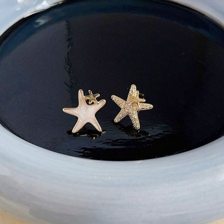 Visão detalhada dos brincos de estrela do mar assimétricos com acabamento em esmalte branco e dourado.