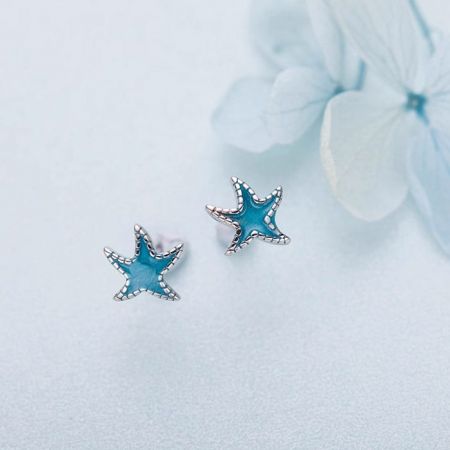 Un paio di piccoli orecchini a bottone a forma di stella marina in smalto blu su una superficie bianca.