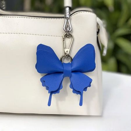Porte-clés et charm en simili cuir avec nœud pour femmes - Un sac à main en cuir blanc élégant orné d'un charm en faux cuir bleu vif en forme de nœud, montrant son utilisation comme accessoire de mode.