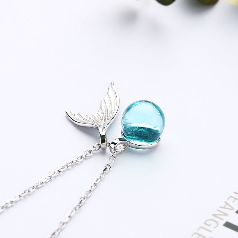 Un primo piano di una collana in argento sterling 925 con un delicato pendente a forma di coda di balena con una perla di luna blu chiaro scintillante.