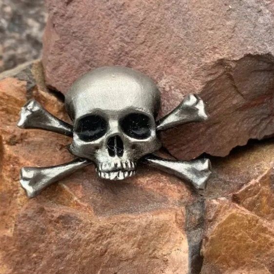 Eine detaillierte Nahaufnahme des Metall-Schädel- und Knochen-Ansteckers aus der Halloween-Kollektion, die das silberfarbene Finish zeigt.