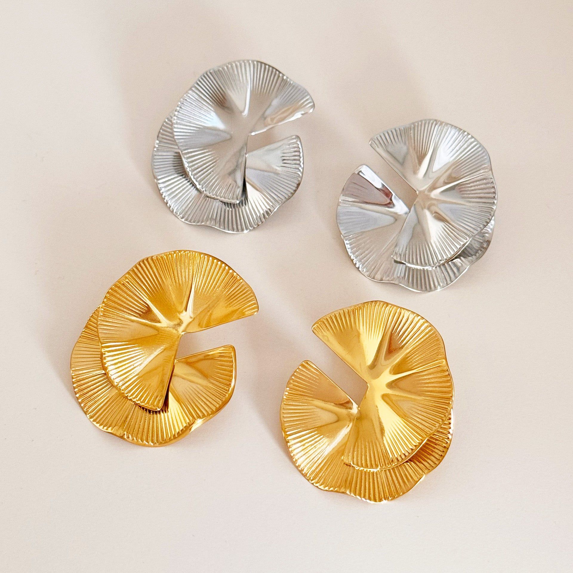 Een set van roestvrijstalen statementoorbellen met twee paar ginkgo blad ontwerpen, één in een gepolijste gouden afwerking en de andere in een strakke zilveren afwerking, weergegeven op een neutrale achtergrond.