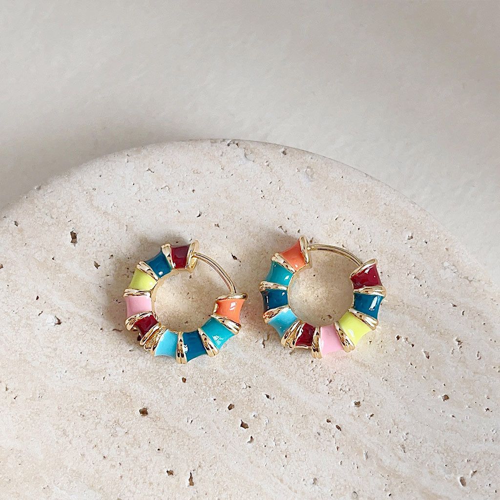 A pair of 14k gold plated bamboo hoop earrings with vibrant rainbow multi-color enamel, displayed on a decorative stone.